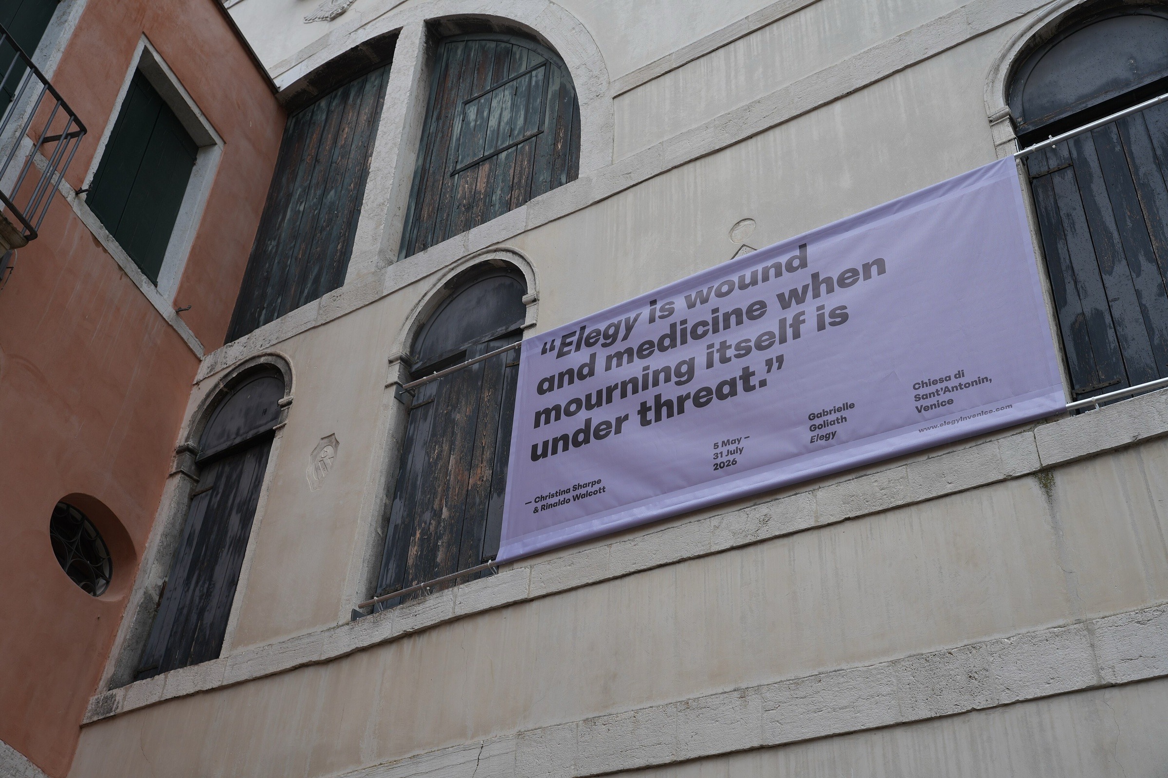 A lilac banner for Elegy hanging on the weathered façade of a Venetian building