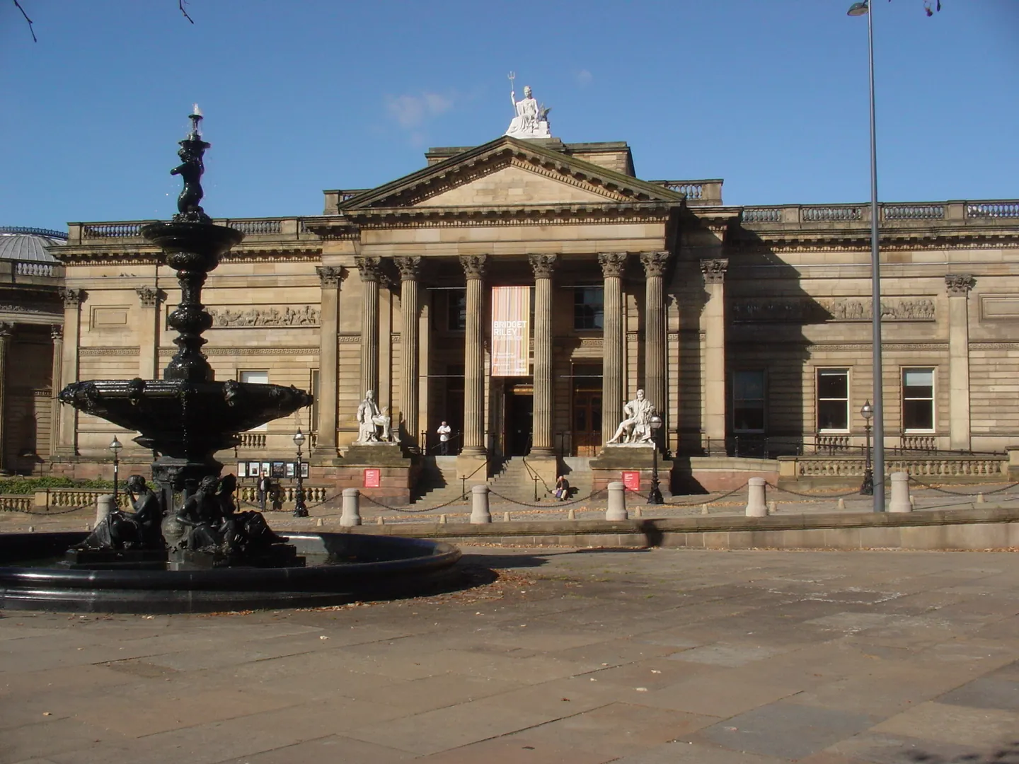 The columned stone front of the Walker Art Gallery in Liverpool, with a Victorian fountain in the foreground under a blue sky