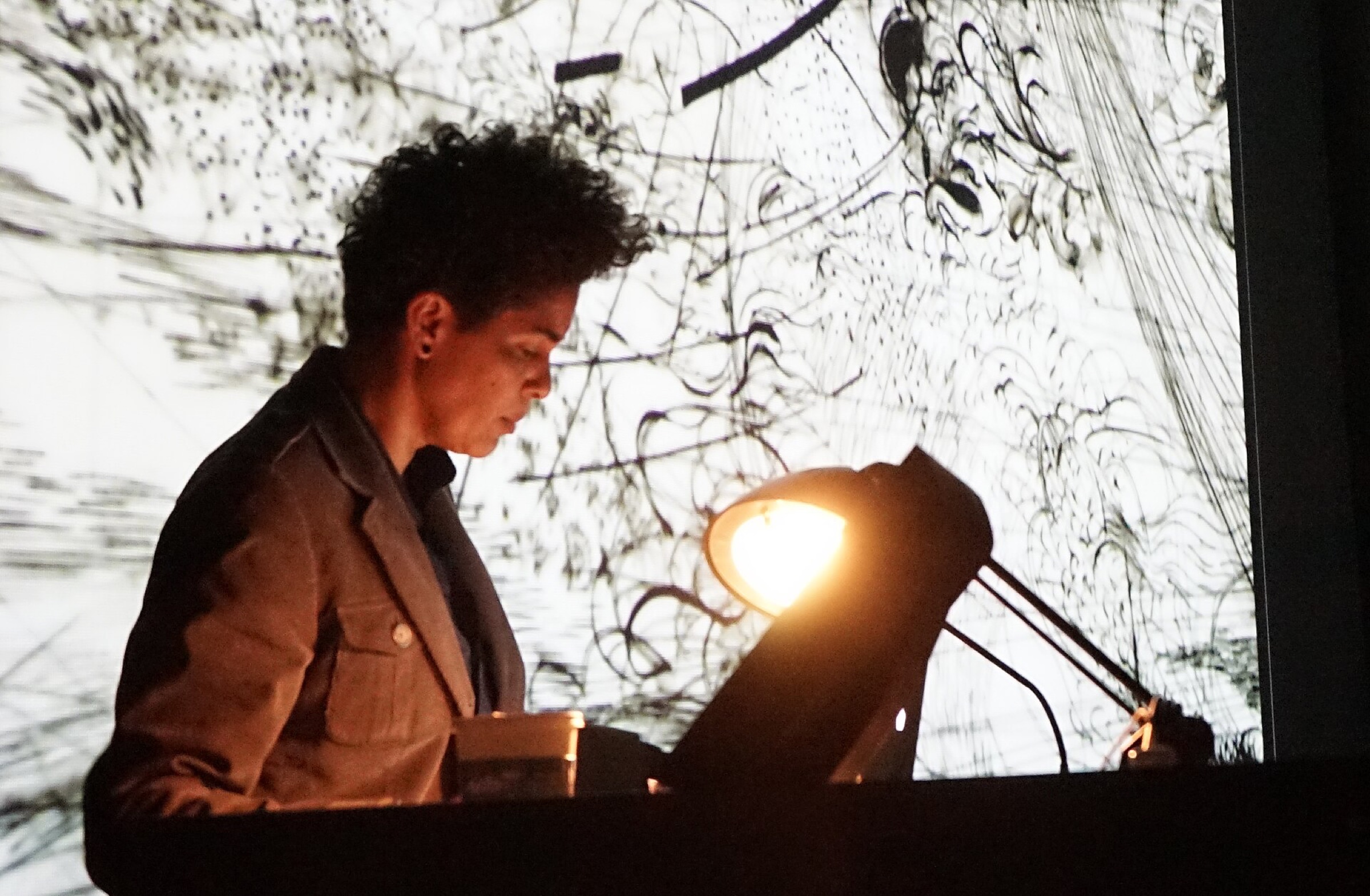 Julie Mehretu at a lectern in front of a projected black and white drawing, lit by a desk lamp