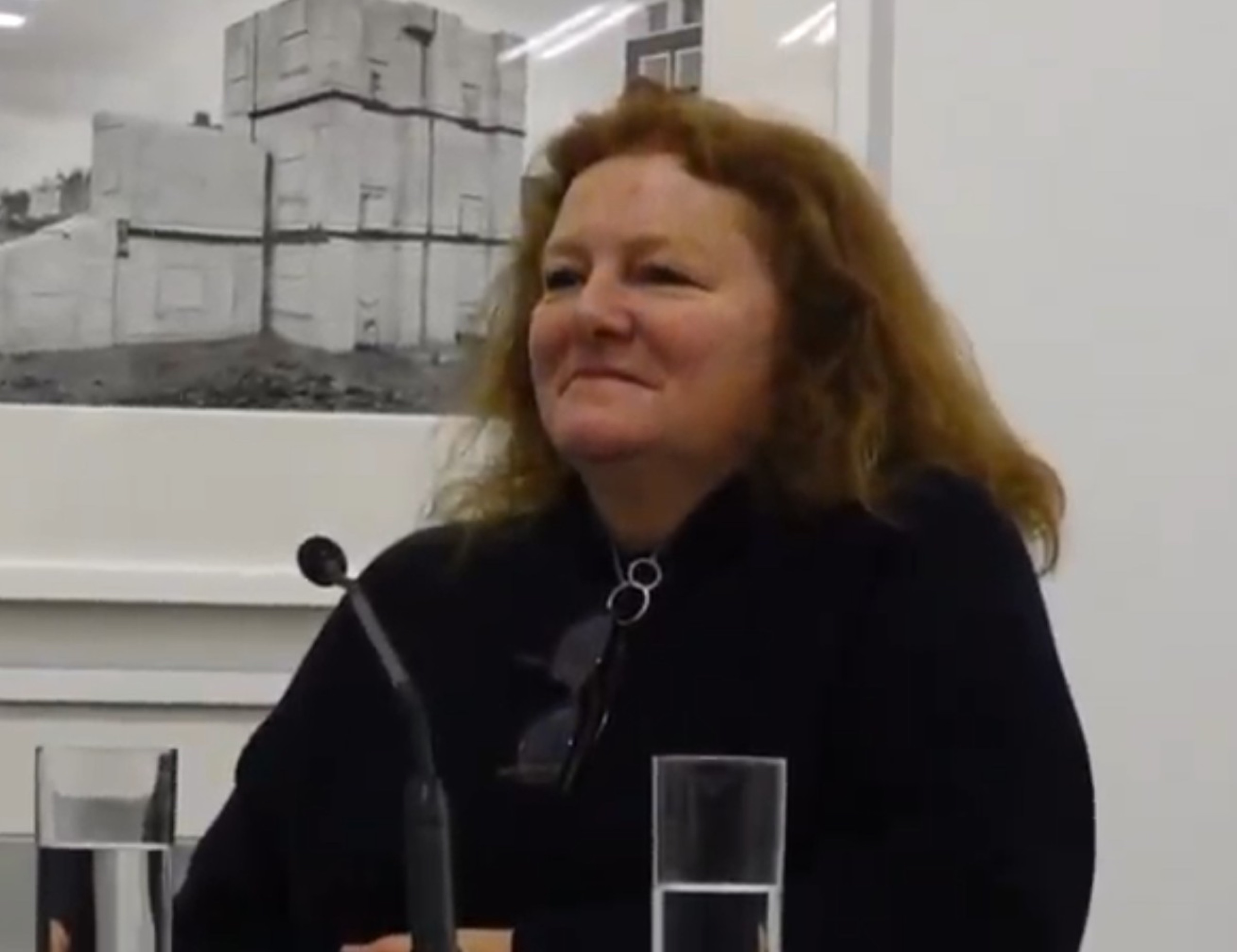 Rachel Whiteread speaking at a press conference, with a photograph of one of her concrete casts on the wall behind her