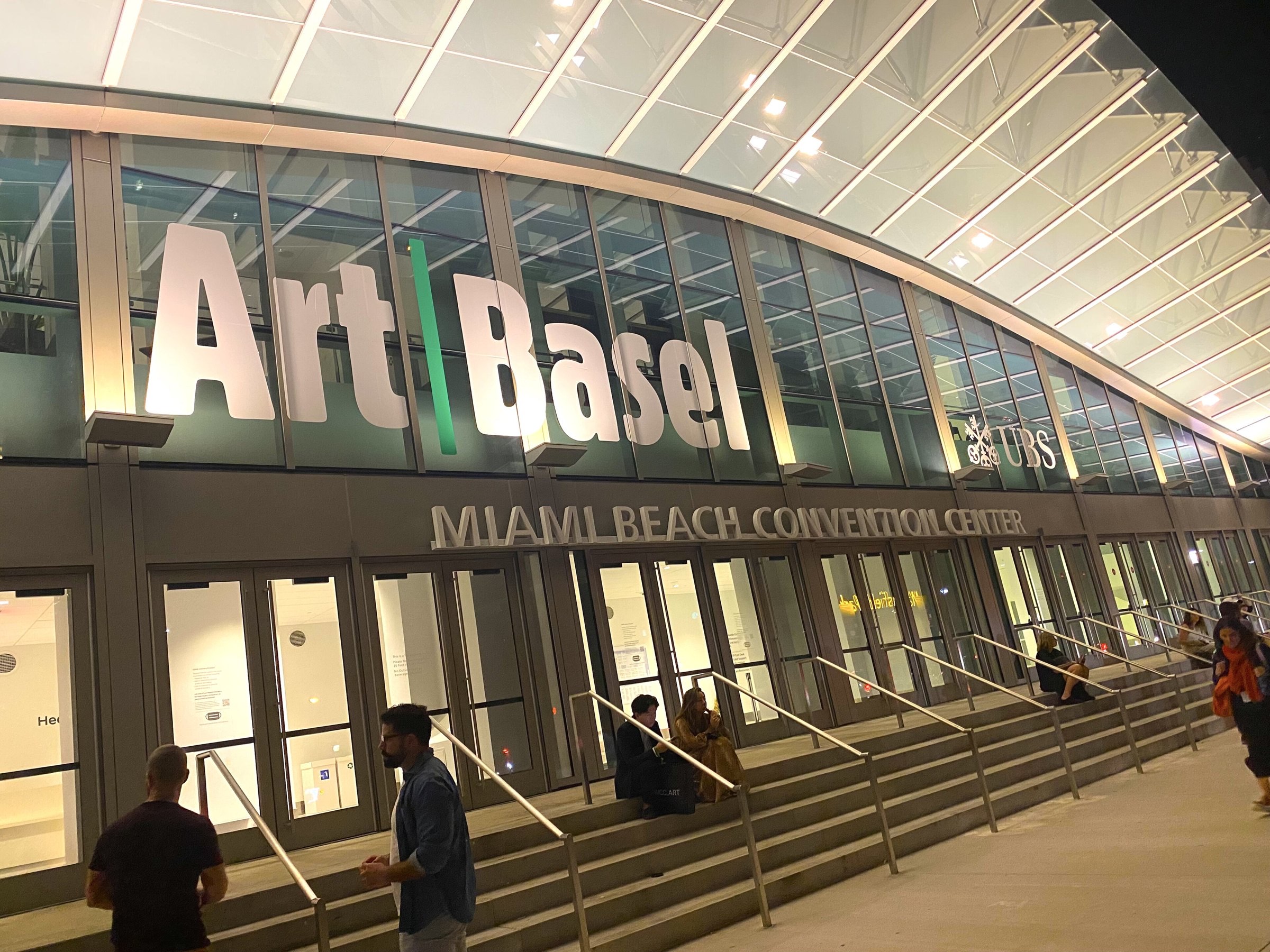The illuminated Art Basel sign above the glass entrance of the Miami Beach Convention Center at night