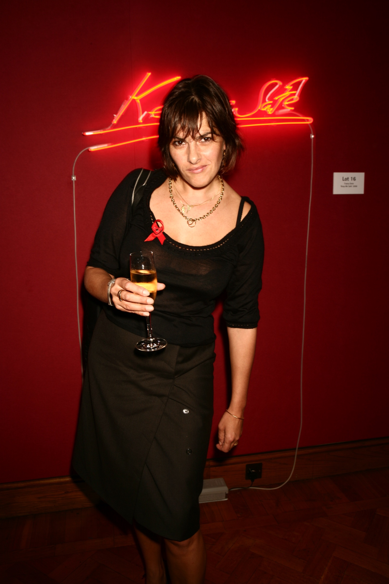 Tracey Emin in a black dress at an evening event, standing in front of a red wall with one of her neon works above her