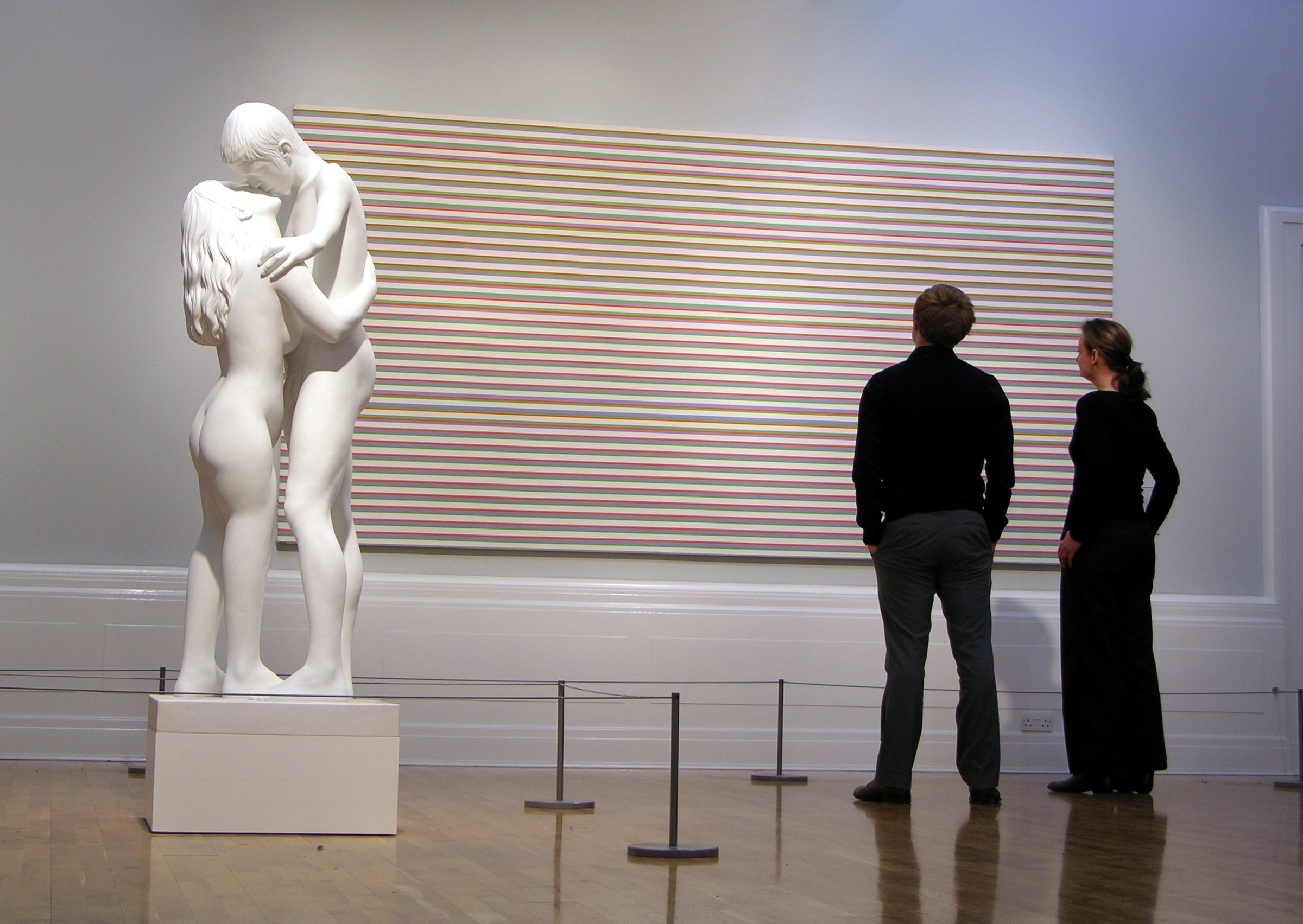 A large painting of fine horizontal stripes in pale colours on a gallery wall, with a white marble sculpture of an embracing couple and two visitors in front of it