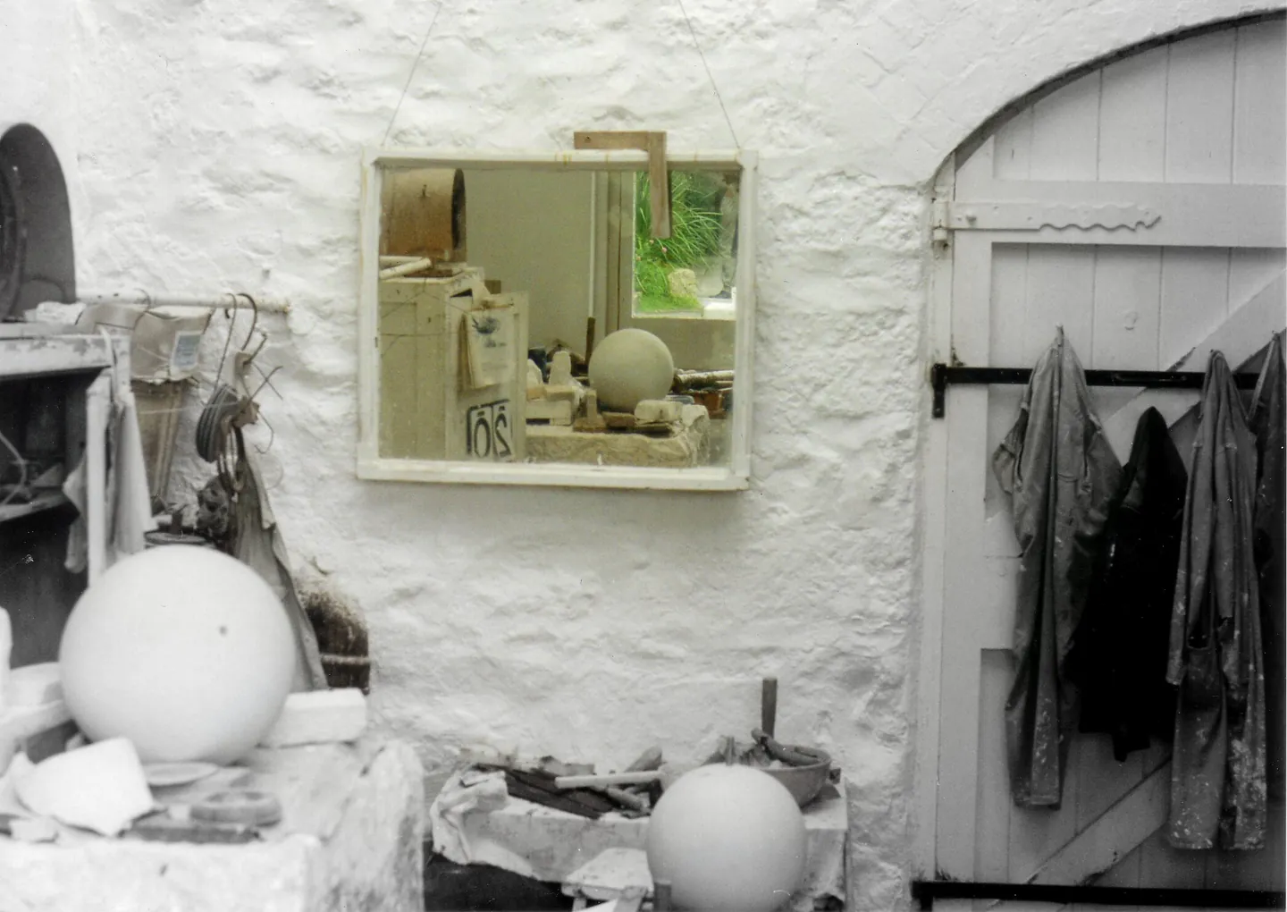 A whitewashed workshop with a wooden door, overalls hanging on hooks, a mirror on the wall and stone spheres on a bench