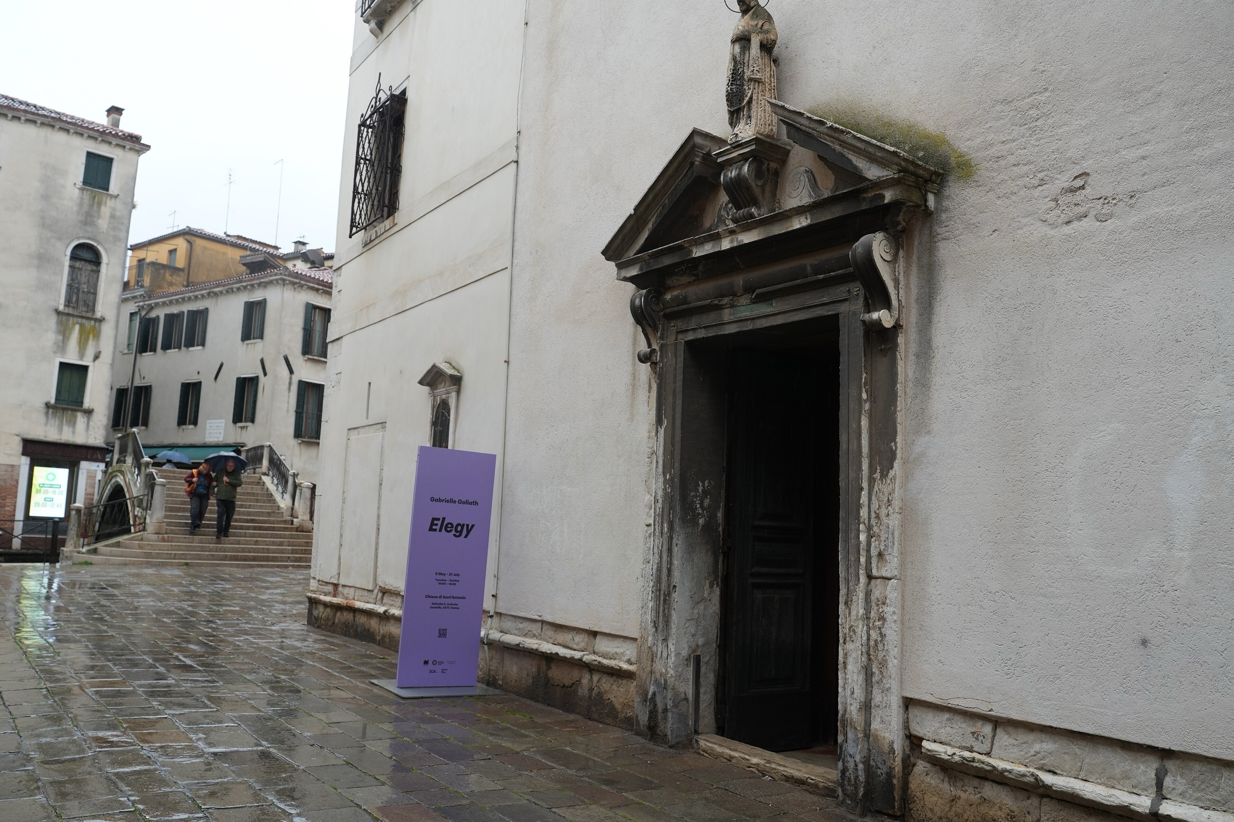 A side door of a white Venetian church with a lilac sign for Elegy beside it, on a wet stone campo