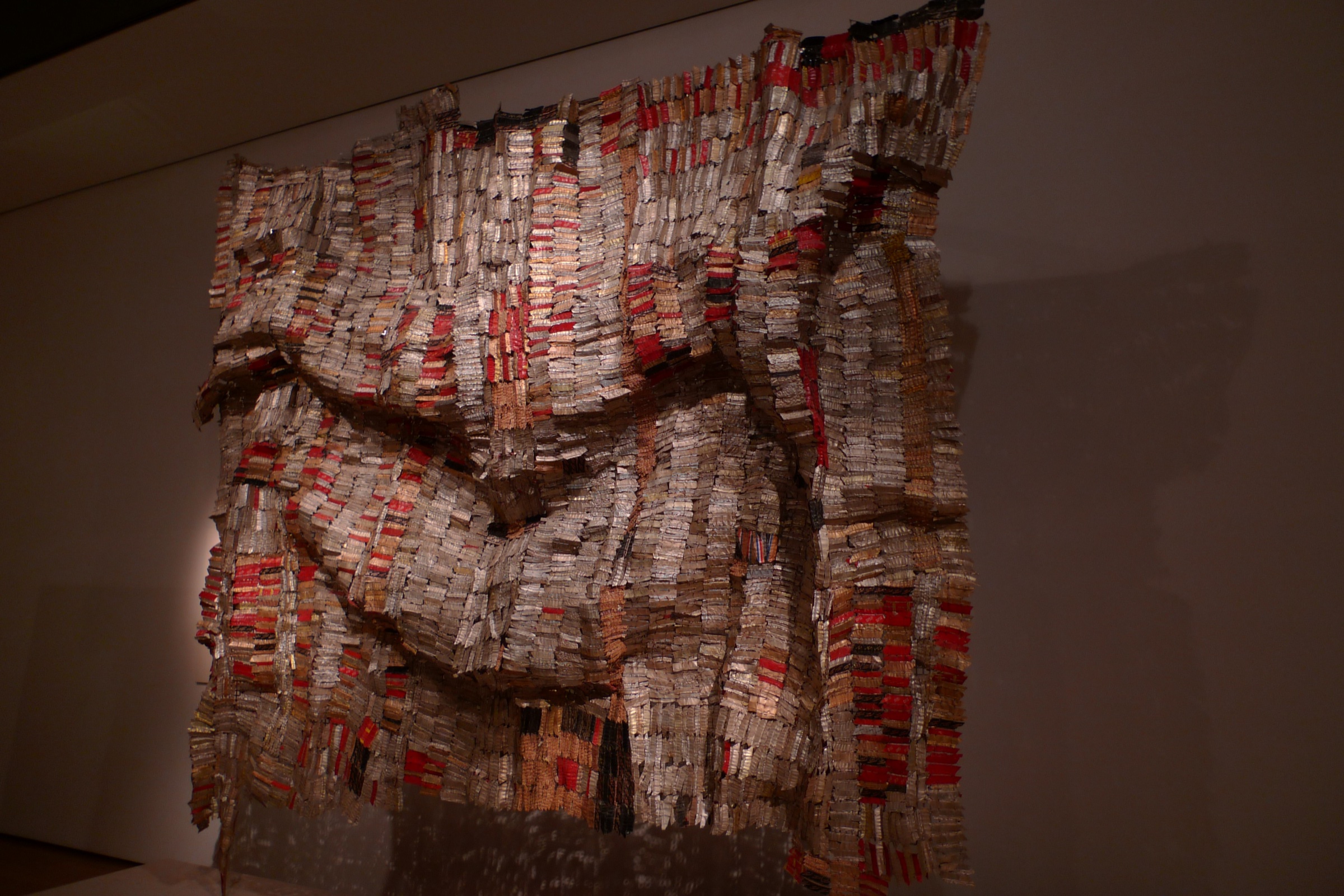 A large hanging made of flattened bottle tops and wire in red, gold and silver, folded like cloth on a gallery wall