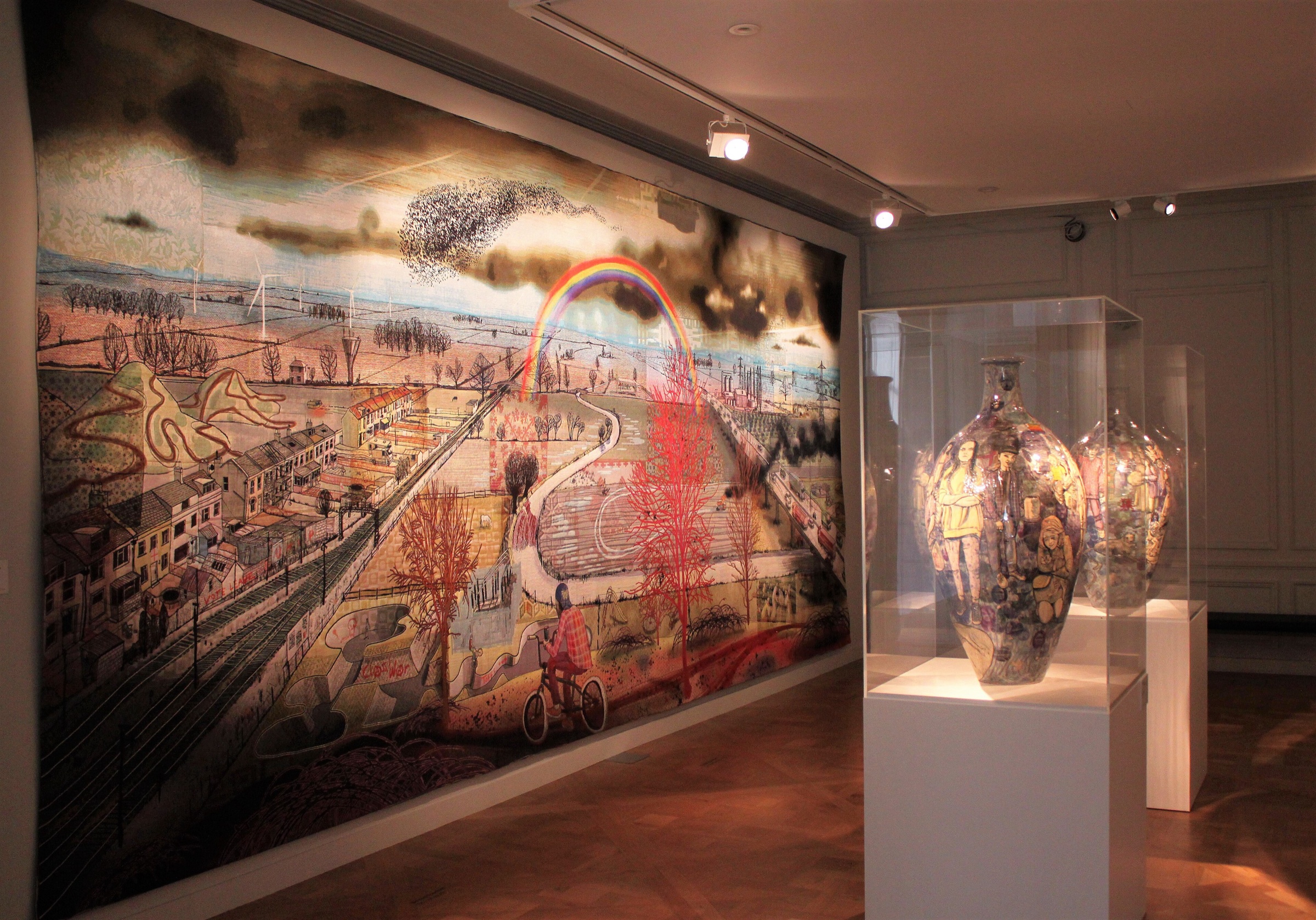 A large Grayson Perry tapestry of a town and a rainbow on a gallery wall, beside two of his vases in a glass case