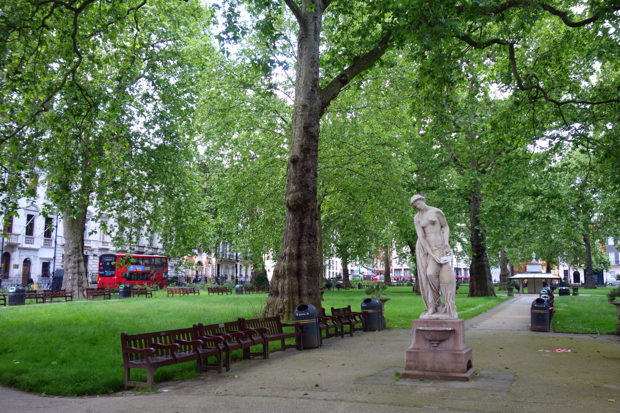 Plane trees and lawns in Berkeley Square, with a white marble statue of a woman on a pedestal beside the path and a red London bus passing beyond the railings