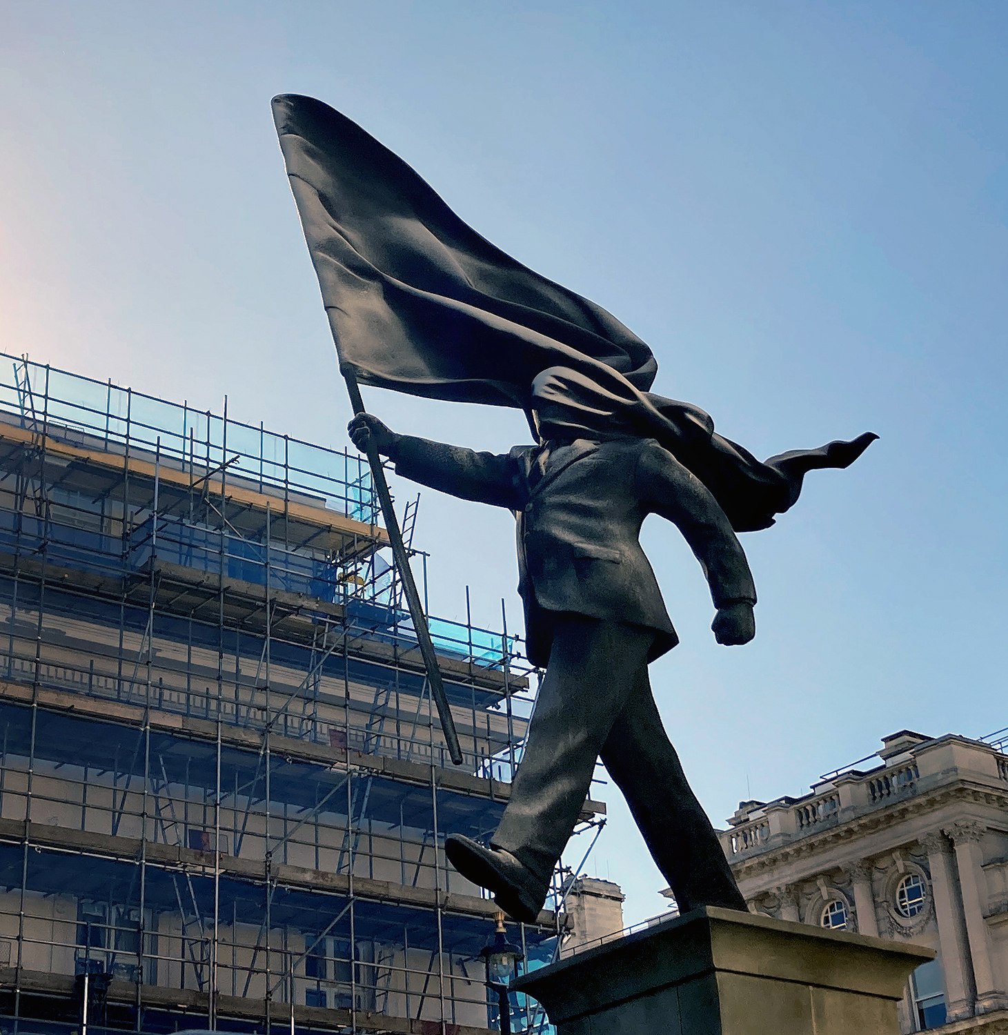 Banksy's Blind Patriotism statue on Waterloo Place, London, in 2026: a bronze-coloured man in a suit strides off his plinth with a flag blowing over his face