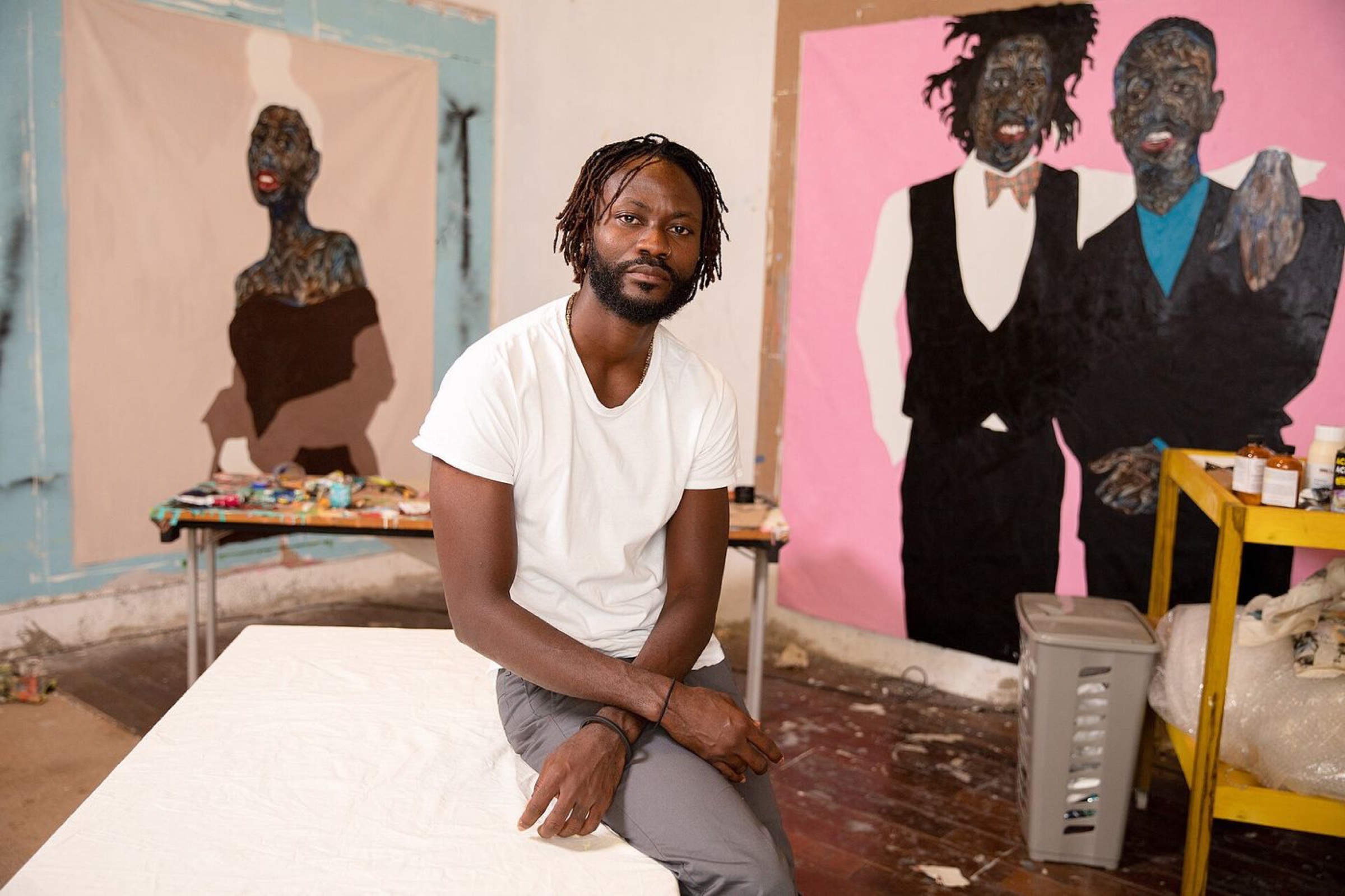Amoako Boafo in a white T-shirt, seated in his studio in front of two of his large portrait paintings
