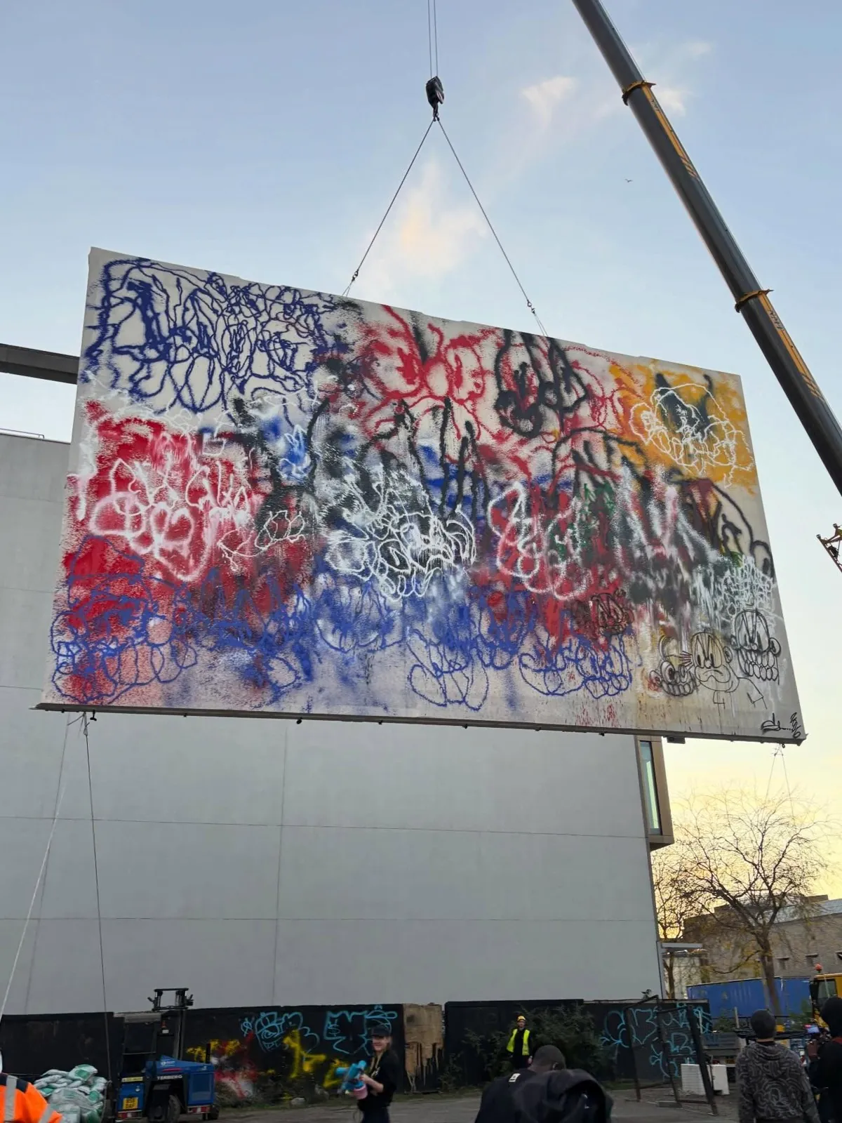 A large Slawn painting in red, blue, black, white and yellow spray paint, covered in scribbled faces and cartoon characters, hanging from a crane above a car park at dusk